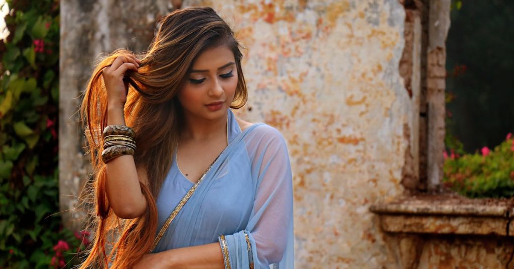 A woman in a blue saree poses gracefully outdoors by a rustic wall.